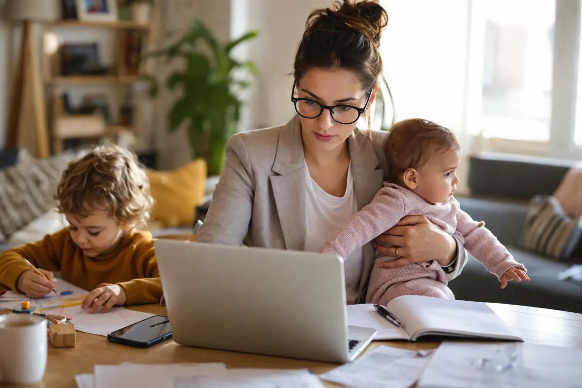 donna che lavora al computer da casa mentre tiene in braccio un bambino e un altro figlio disegna accanto